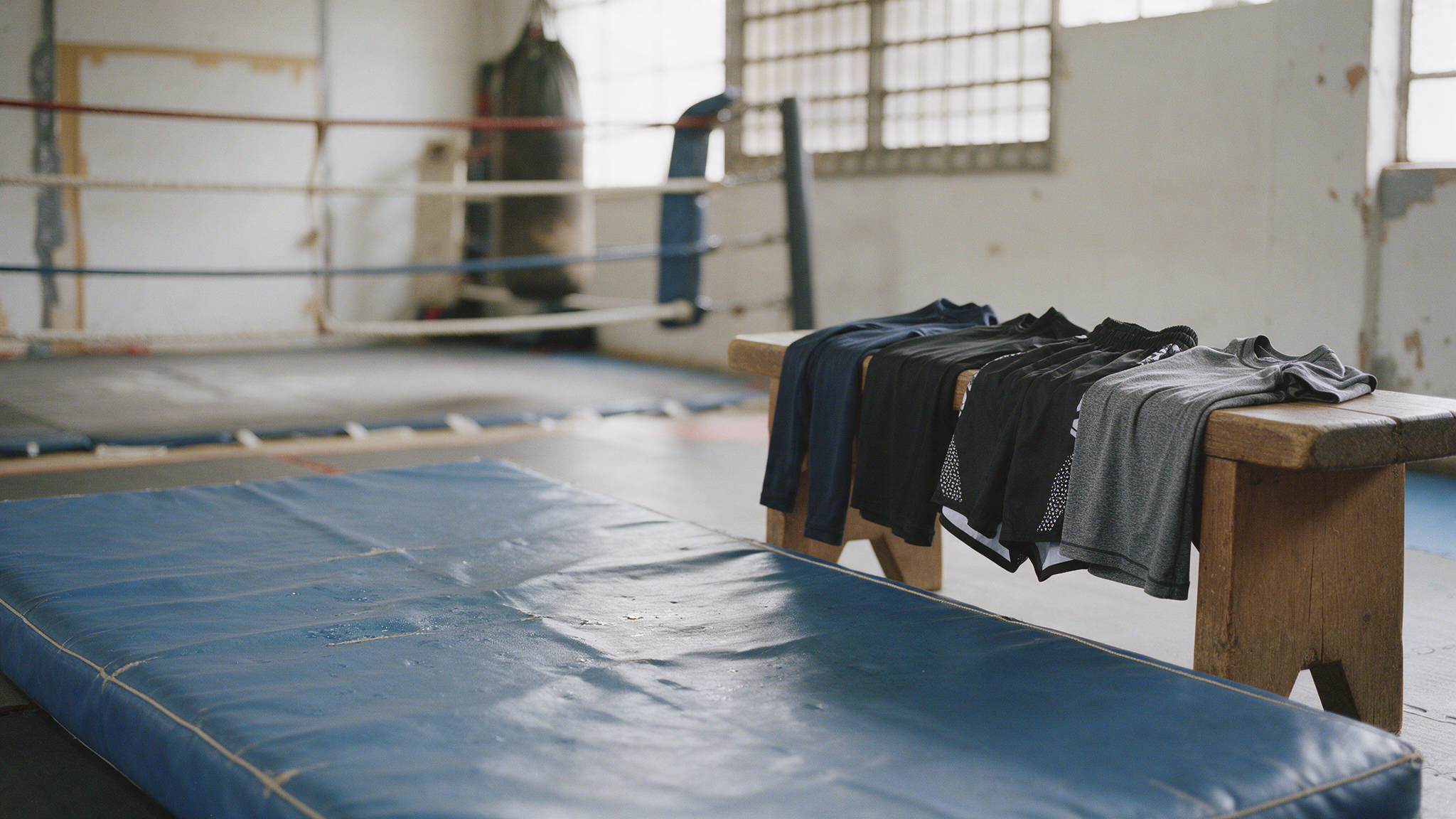 Rashguards et fight shorts pliés sur un banc dans une salle de sports de combat, ring de boxe et sac de frappe en arrière-plan
